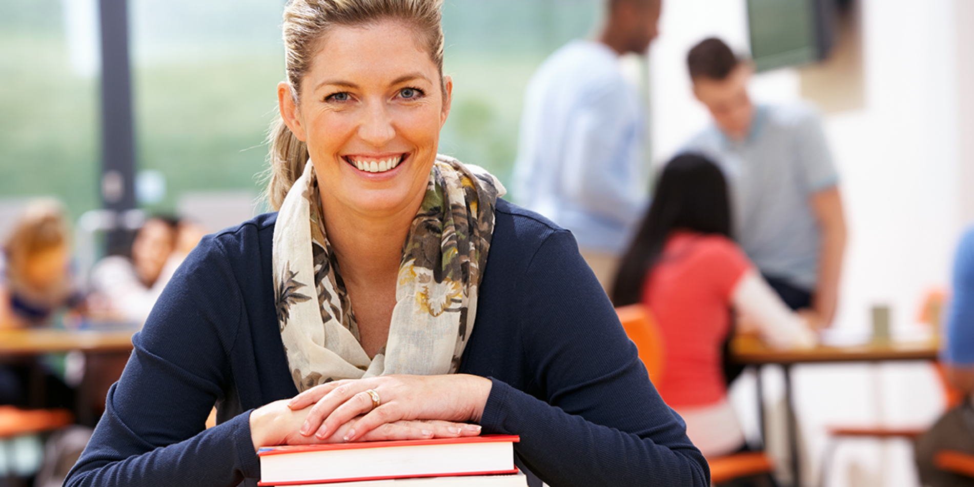 Mature female student smiling with hands on stack of books, in classroom with other students